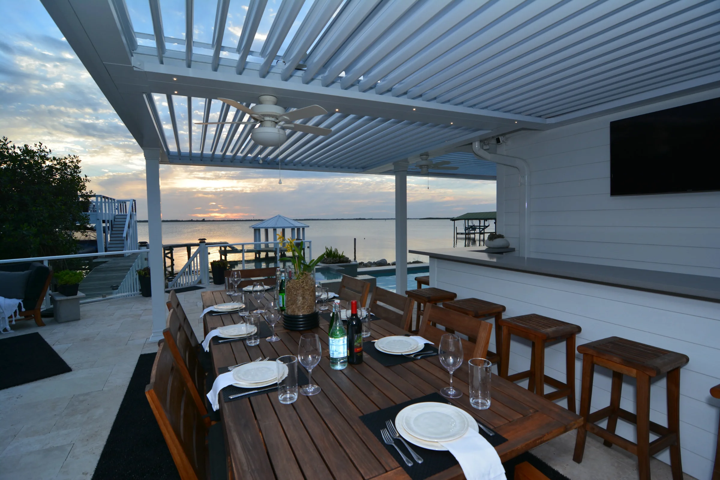 Louvered aluminum pergola over a waterfront dining terrace at sunset with open water beyond