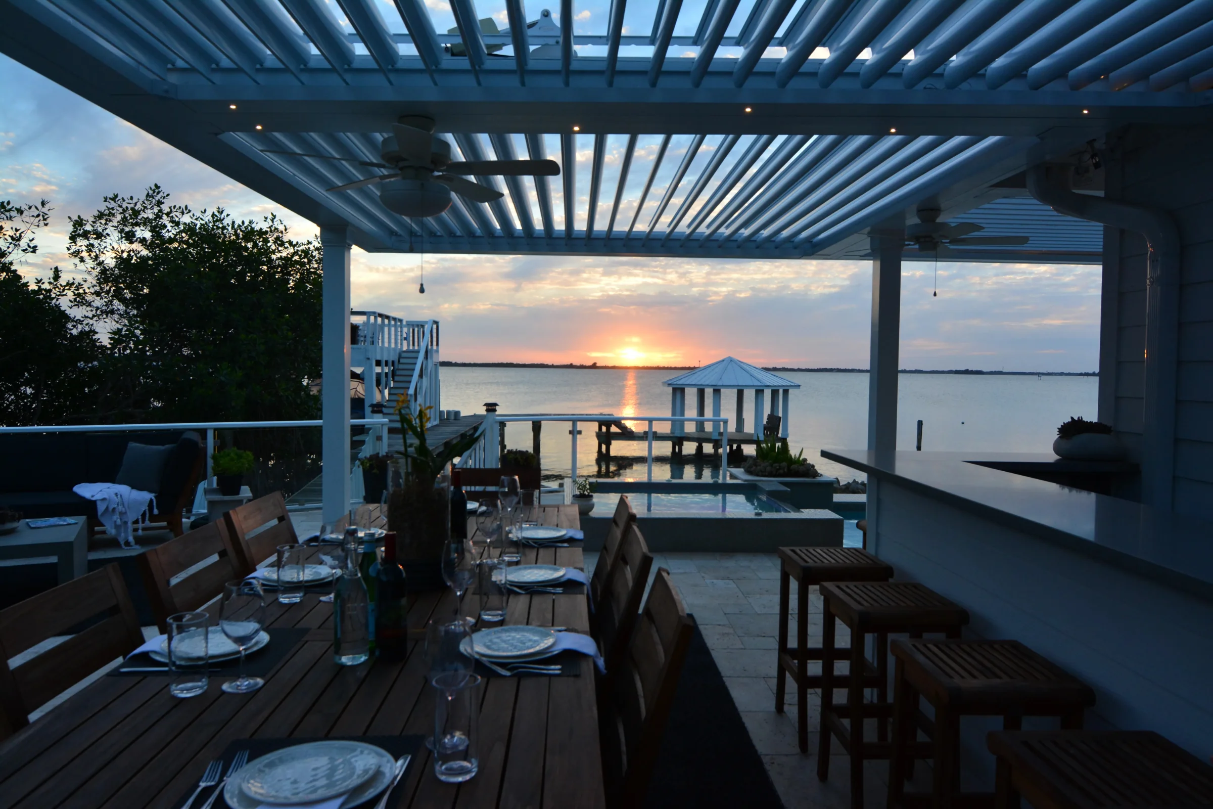 Waterfront dining area under an aluminum louvered pergola with warm sunset light over the water