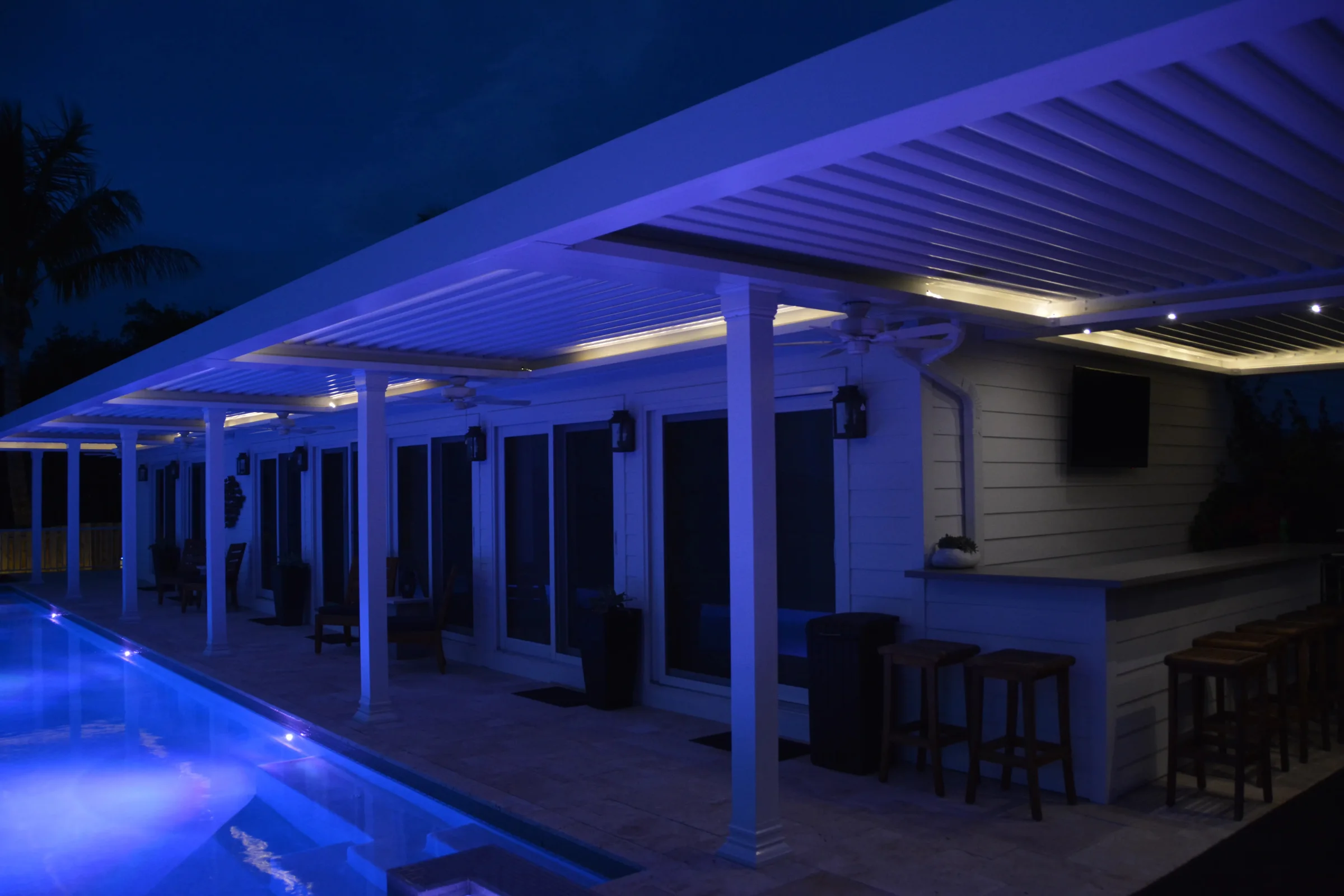 Aluminum louvered pergola over a pool patio at night with blue accent lighting and an outdoor bar