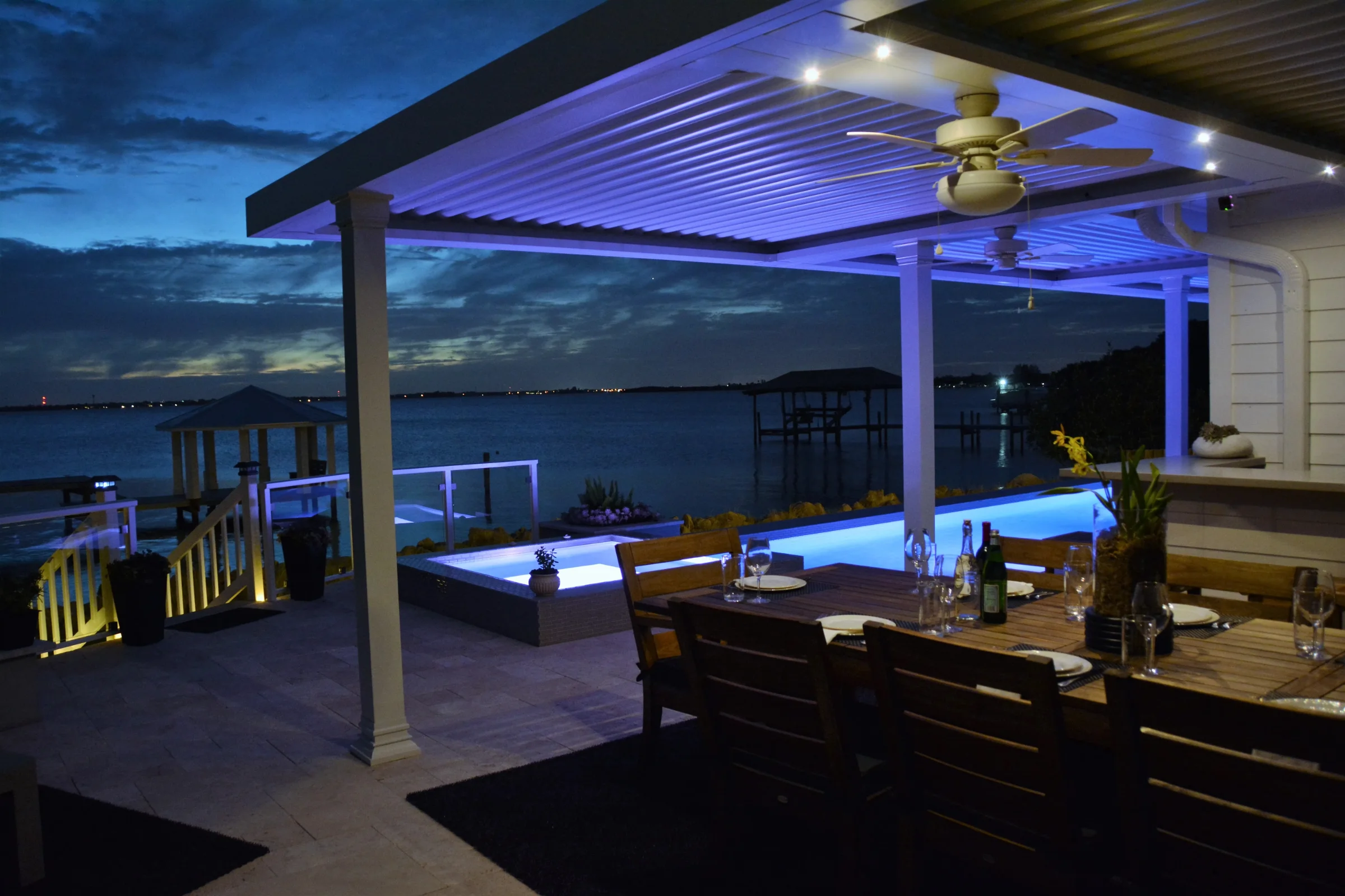 Louvered pergola lit at night over a waterfront patio with a dock and open water behind it