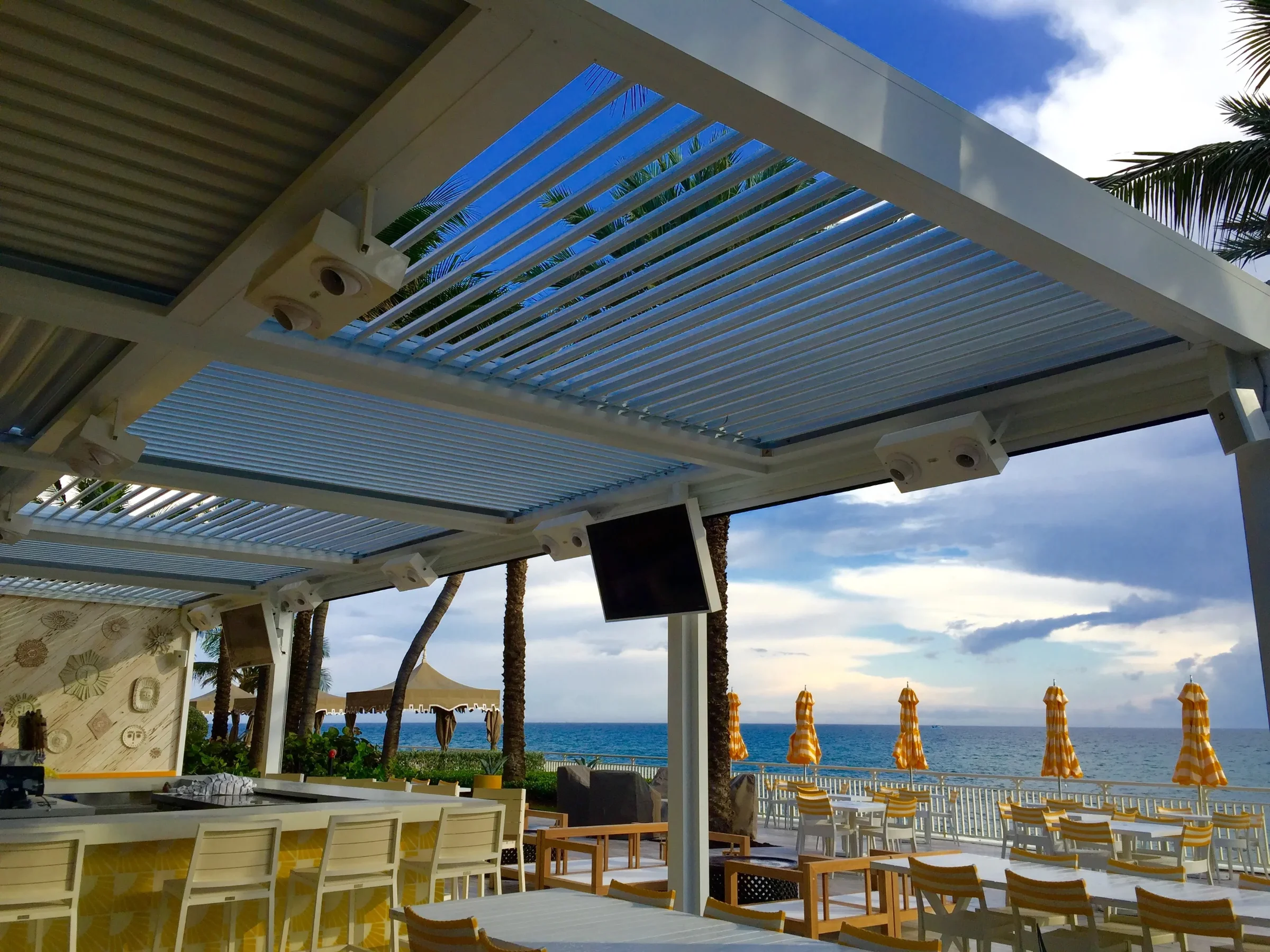 Oceanfront dining terrace shaded by a white aluminum louvered pergola with a bar beyond