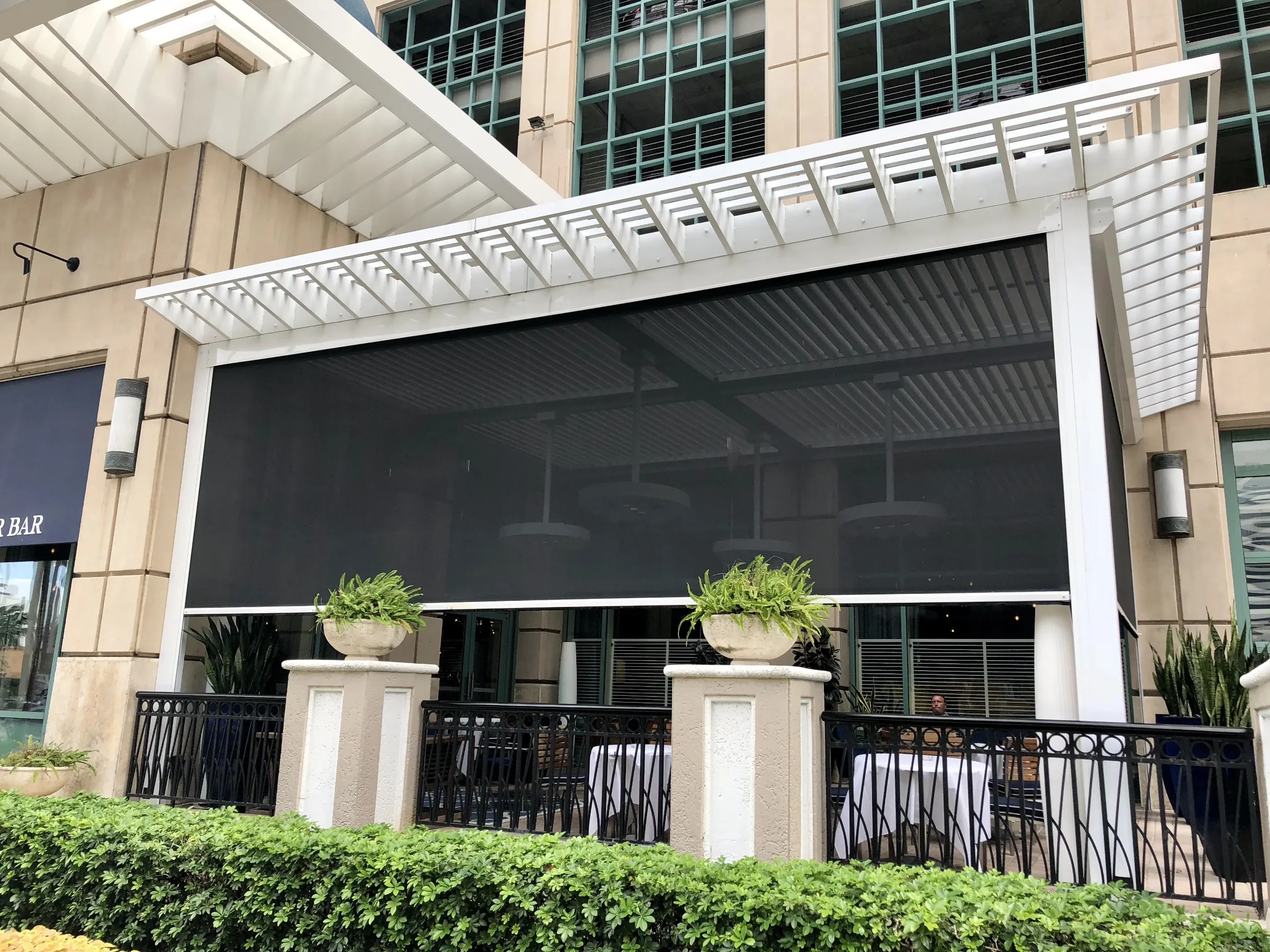 Dark motorized screen fully deployed on a commercial patio beneath a white louvered pergola roof