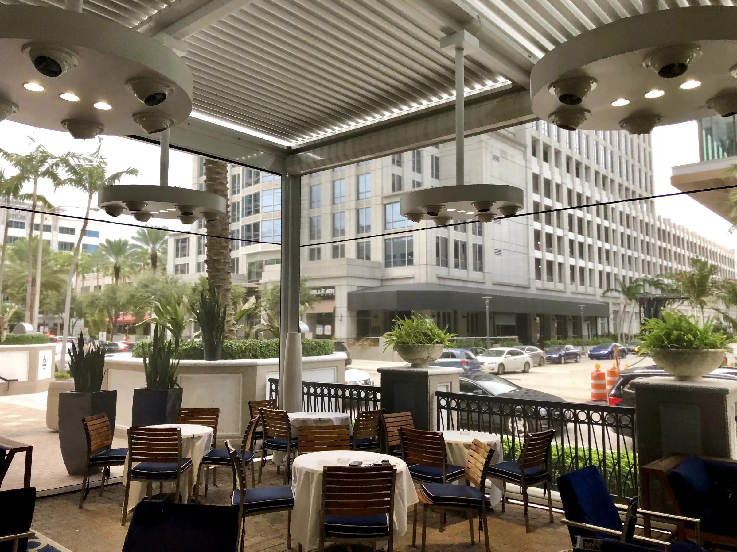Commercial outdoor dining terrace with a white louvered pergola roof, pendant lighting and city buildings behind