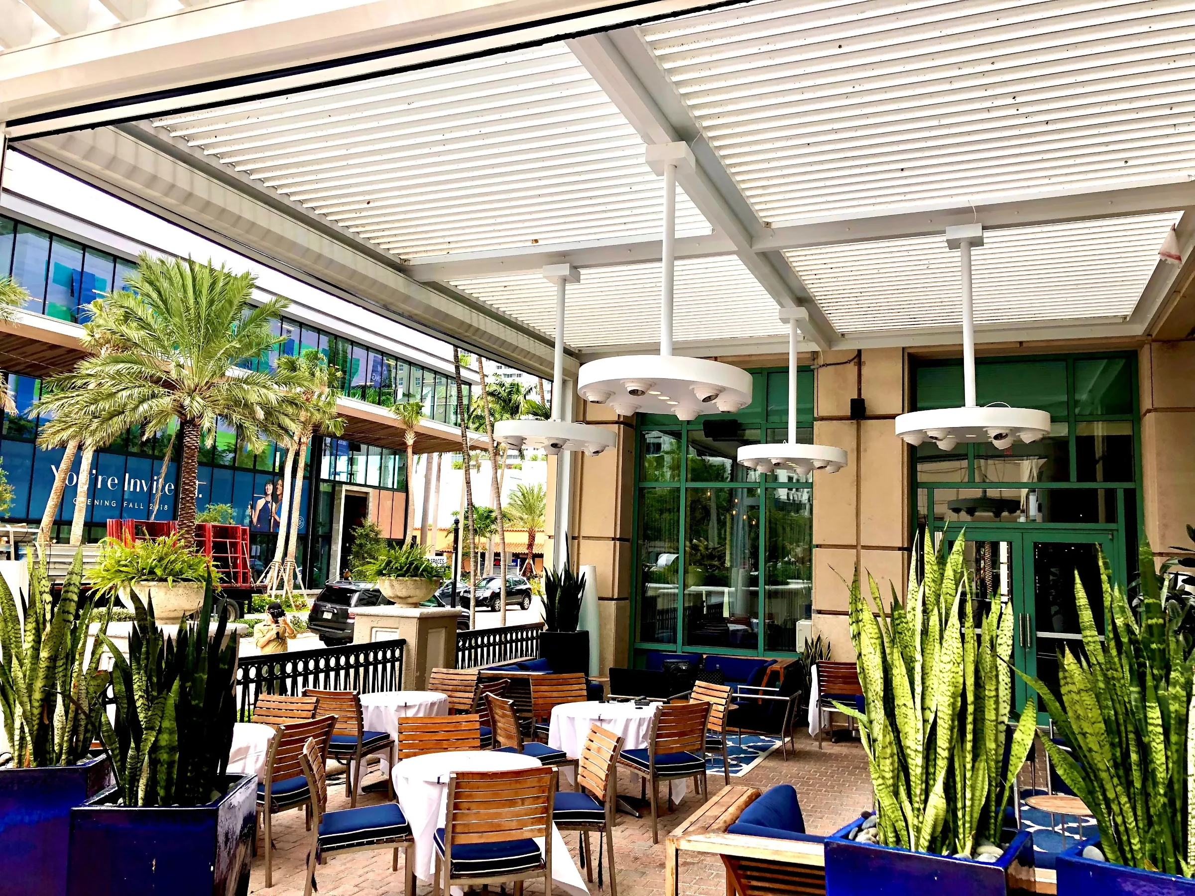 White commercial louvered pergola over a restaurant patio with palm trees and blue umbrellas